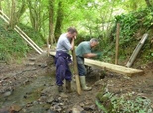 Photograph showing two men using timber to make a footbridge across a small river in the woodland area on Kilkhampton Common