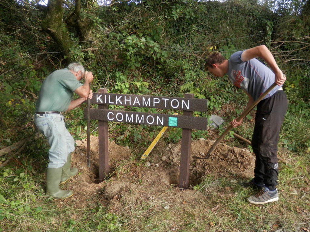 Two men are shovelling soil into post holes for the new "Kilkhampton Common" sign.