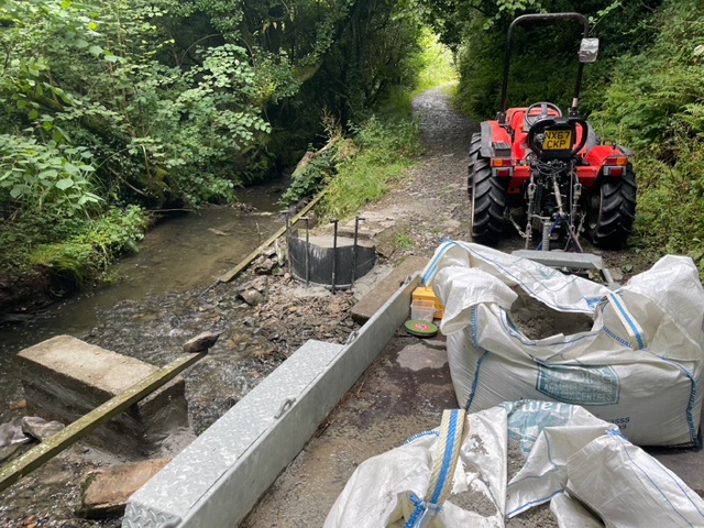 Concrete piers set into a stream in a wooded area. A tractor and building materials for the piers construction are to the right of the image in dumpy bags.